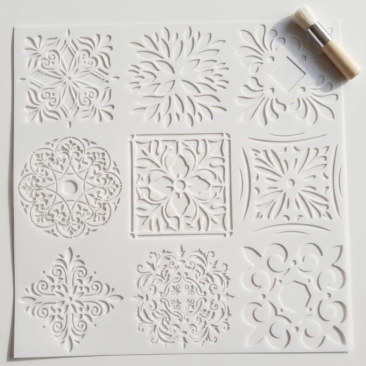 White square 2020-45X45-3 paint stencil featuring nine intricate ornamental patterns, accompanied by a small brush.