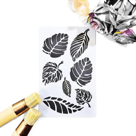 White reusable plastic stencil sheet for painting with assorted detailed leaf cutouts, displayed on a dark surface.