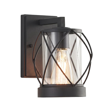 Black metal exterior wall light with clear glass cylinder and geometric cage, featuring an exposed Edison bulb, viewed from