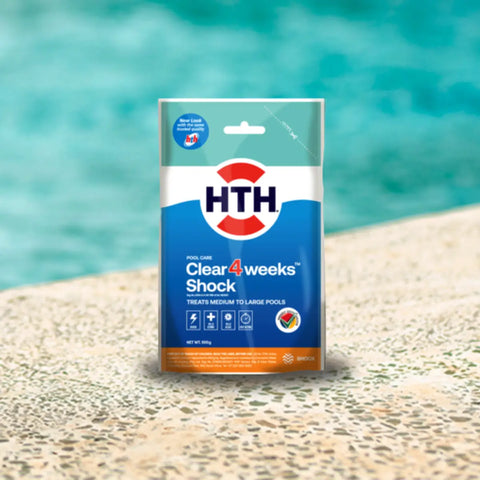 HTH pool care four weeks shock product in blue and white packaging sits on sandy poolside with turquoise water behind