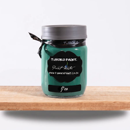 J'Co 250ml Tjohoko Paint master chalk paint in a teal color glass jar with a dark lid and ribbon, sitting on a wooden shelf.