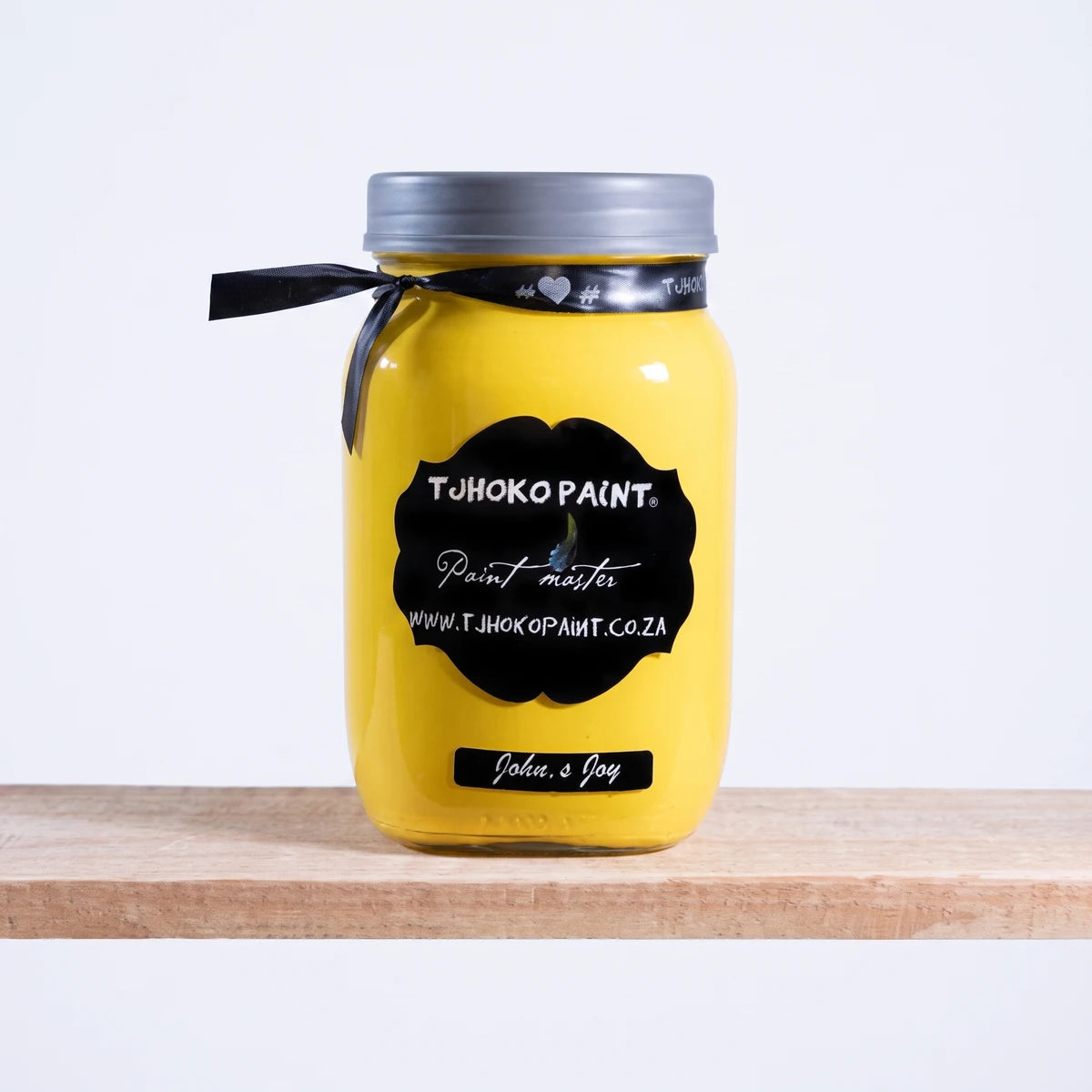 John's Joy 1L Chalk Paint in a vibrant yellow glass jar with a black label and ribbon, resting on a light wooden shelf.