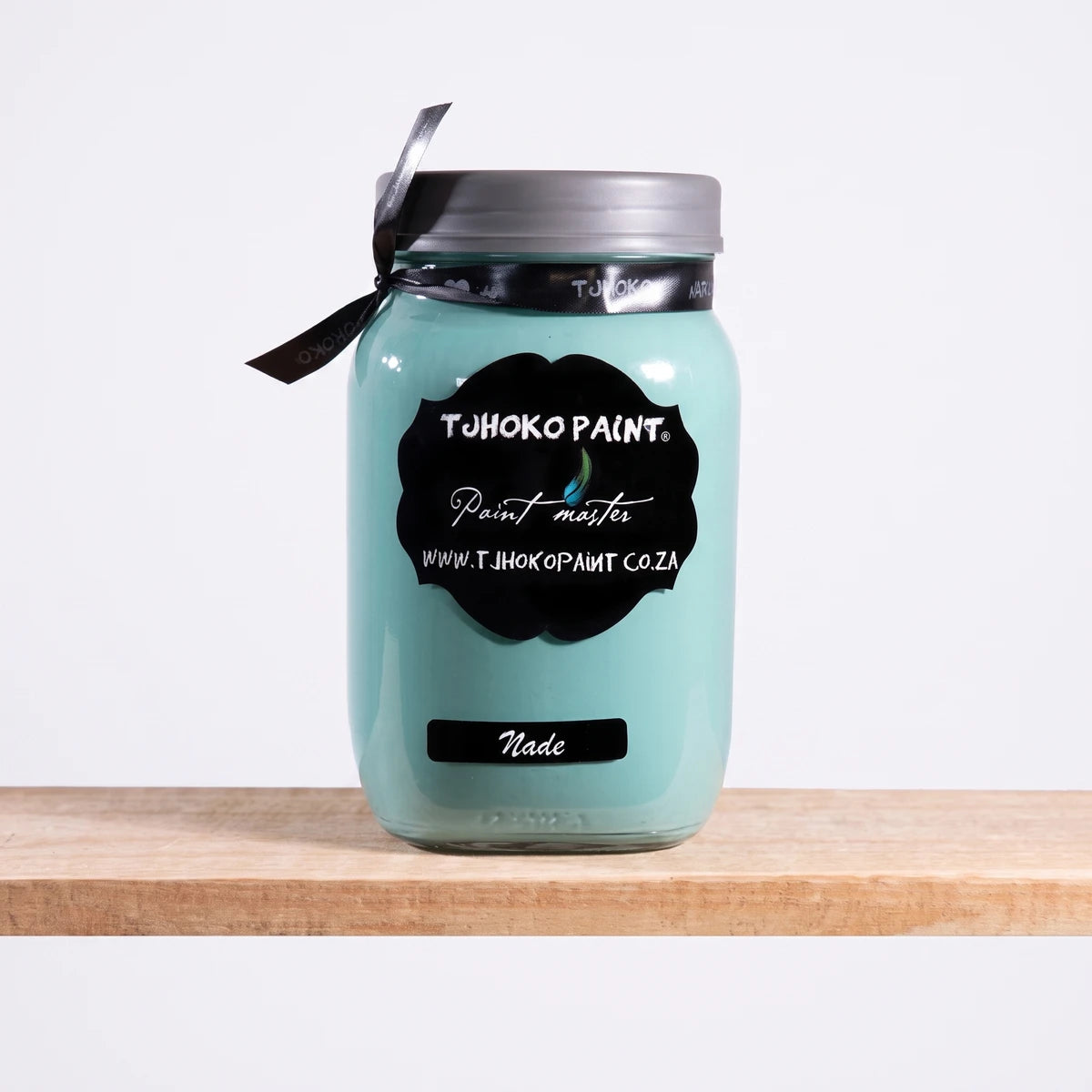 A glass jar of TJ Hoko Paint in Nade, a light teal chalk paint, with a black label, displayed on a wooden shelf.