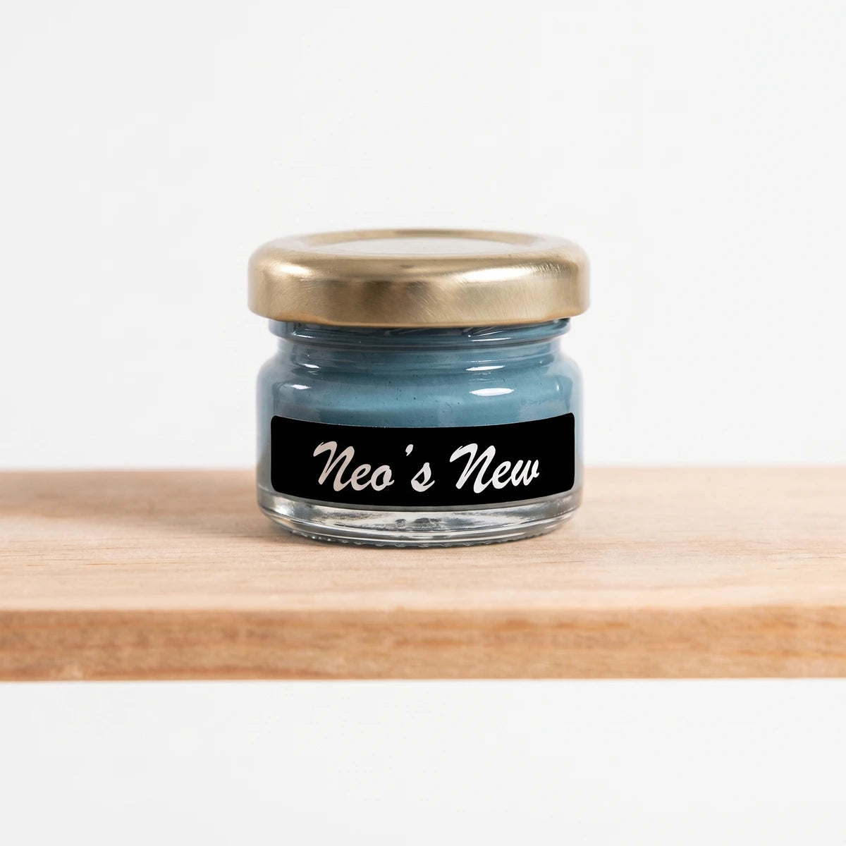 Small glass jar of Neo's New 25ml blue chalk paint with a golden lid and black label, resting on a wooden shelf.