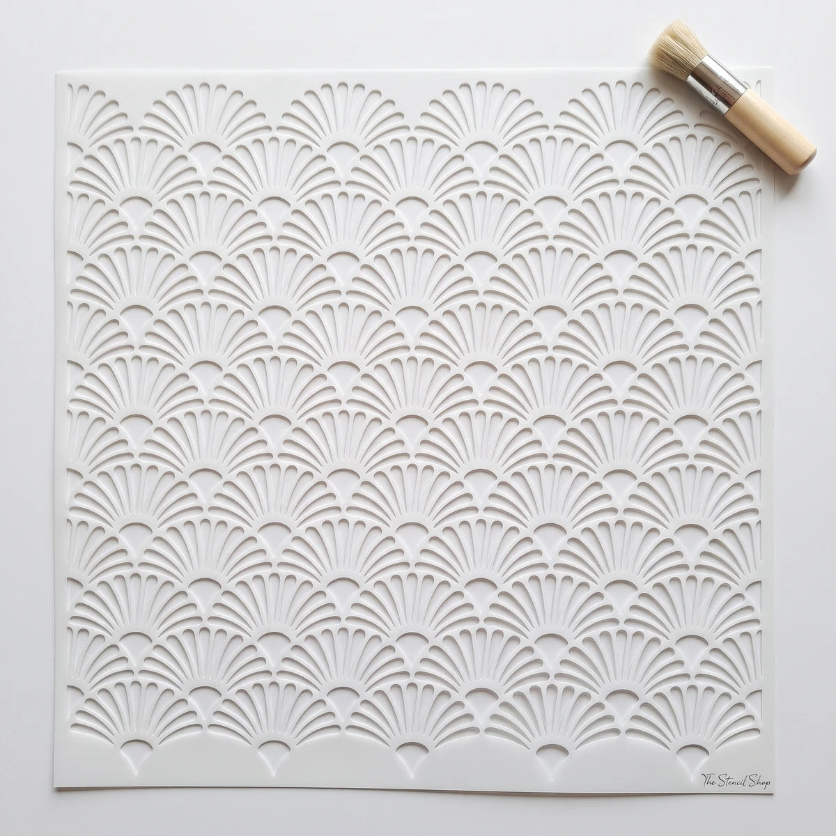 White paint stencil with an intricate Art Deco fan pattern, shown with a small natural bristle brush.