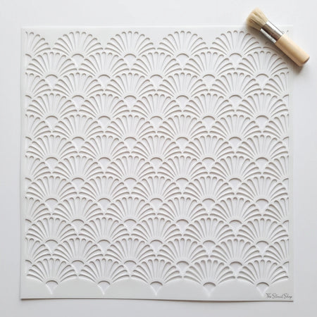 White paint stencil with an intricate Art Deco fan pattern, shown with a small natural bristle brush.