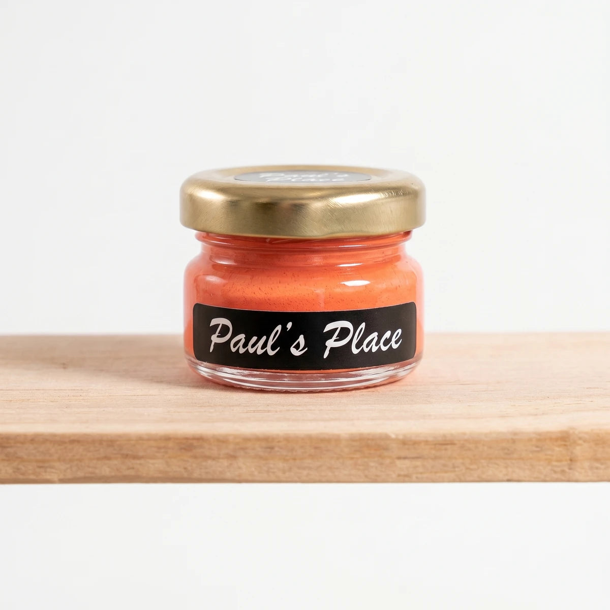 Small glass jar of Paul's Place 25ml orange chalk paint with a gold lid and black label, standing on a wooden shelf.