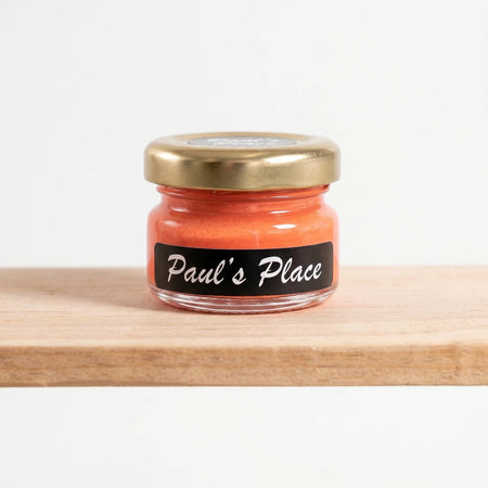 Small glass jar of Paul's Place 25ml orange chalk paint with a gold lid and black label, standing on a wooden shelf.