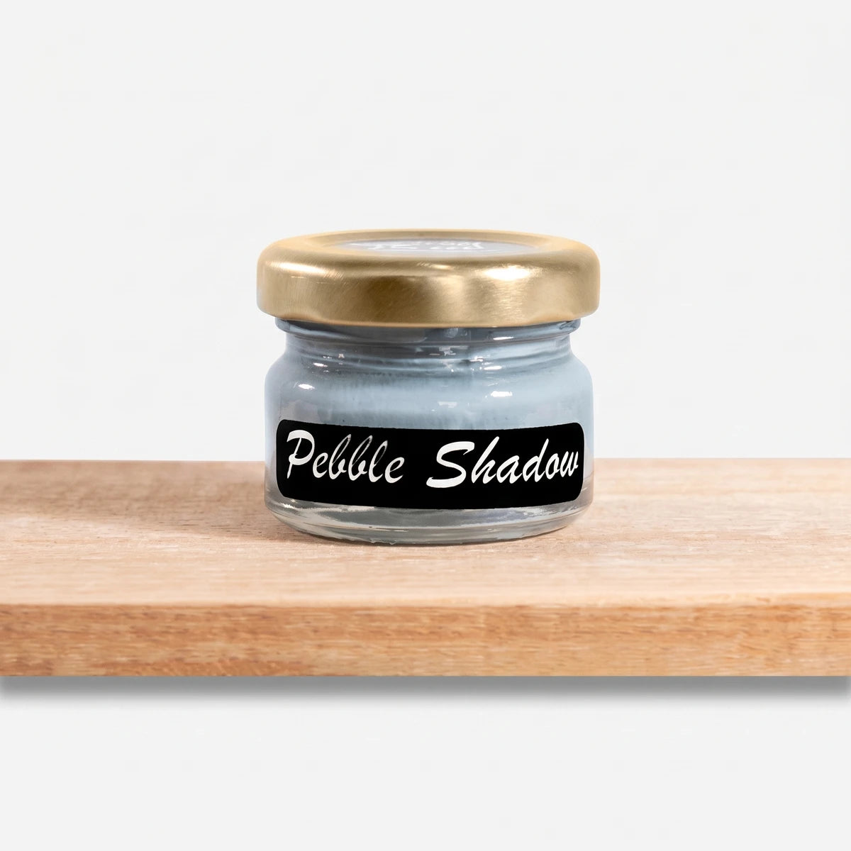 Pebble Shadow 25ml chalk paint in a glass jar with a gold lid, showcasing its light blue-grey hue on a wooden shelf.