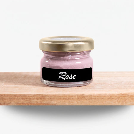A small clear glass jar filled with rose pink chalk paint, featuring a gold lid and a black label with 'Rose' written in
