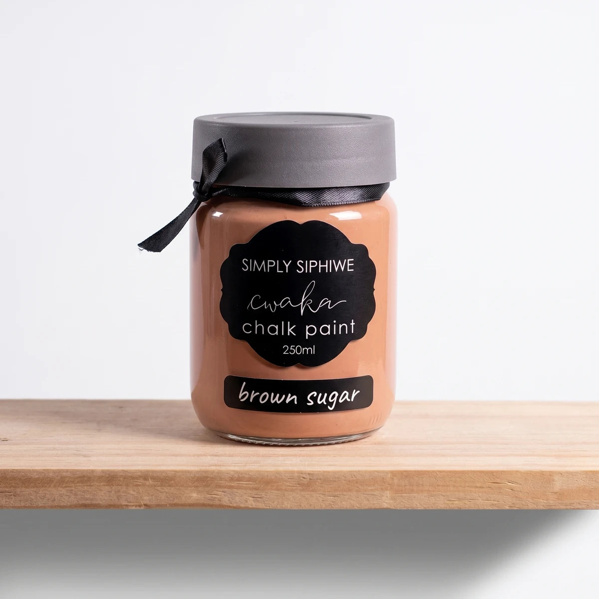 Simply Siphiwe 250ml Chalk Paint jar in brown sugar color, with a black lid and ribbon, on a wooden shelf.