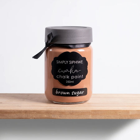 Simply Siphiwe 250ml Chalk Paint jar in brown sugar color, with a black lid and ribbon, on a wooden shelf.
