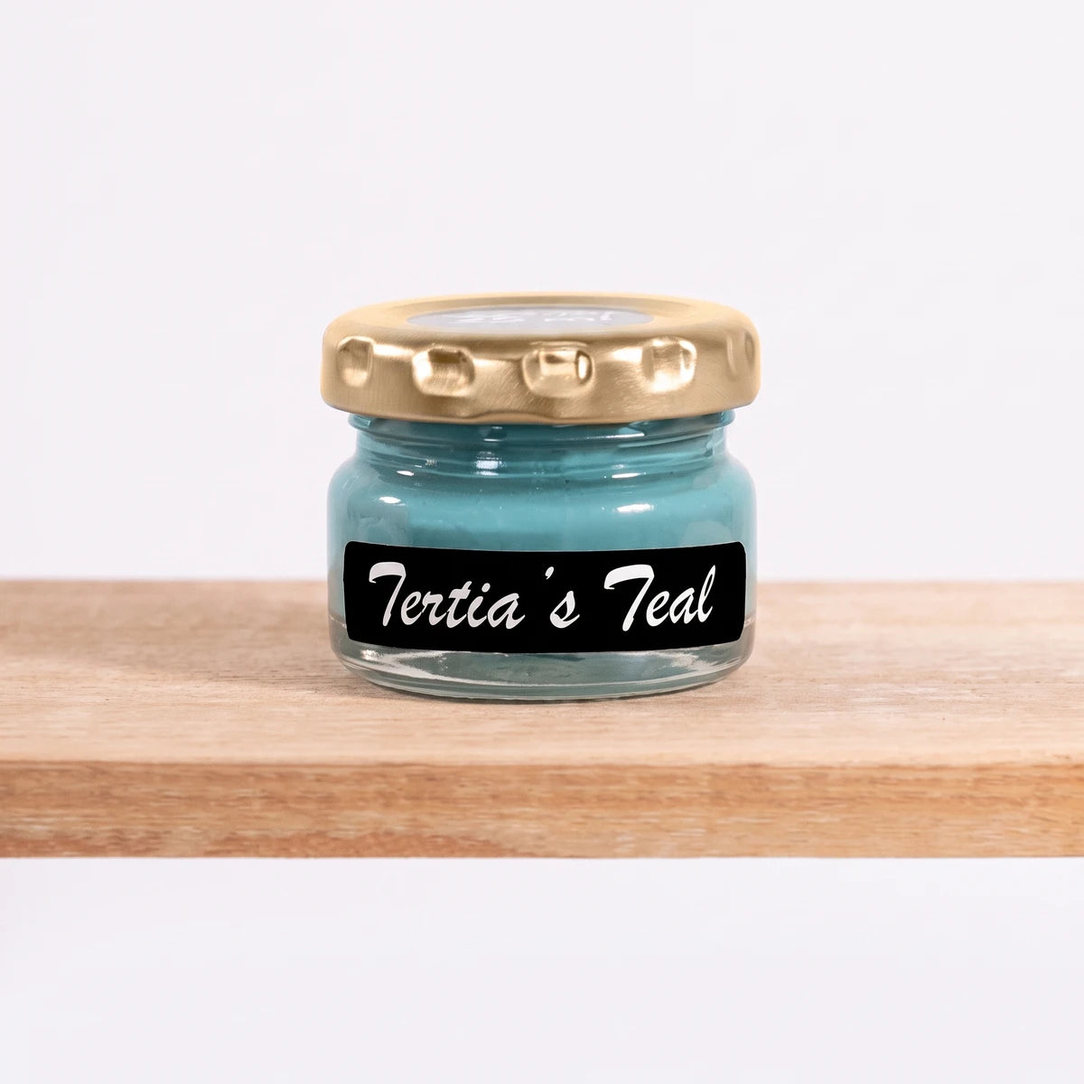 Tertia's Teal 25ml chalk paint in a small clear glass jar with a gold lid and a black label, on a wooden shelf.