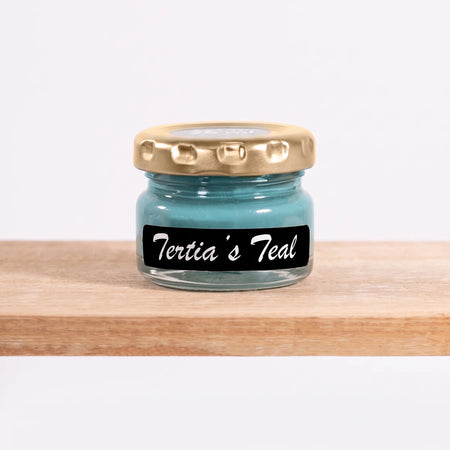 Tertia's Teal 25ml chalk paint in a small clear glass jar with a gold lid and a black label, on a wooden shelf.