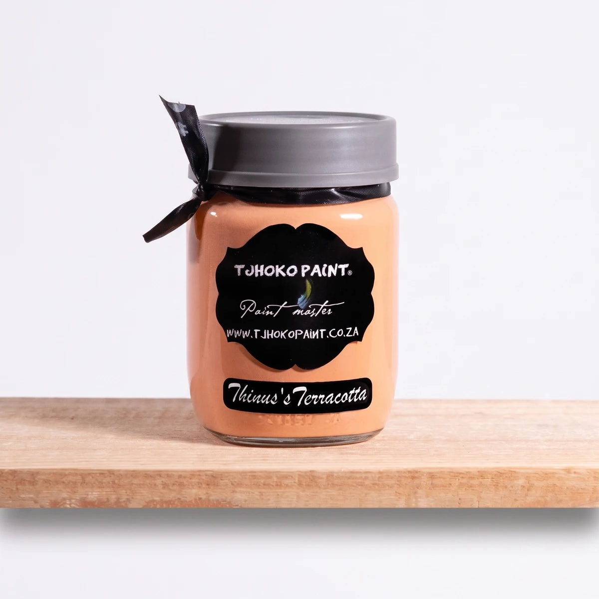 Thinus's Terracotta 250ml Tjohoko Chalk Paint in a clear glass jar with a black label and ribbon, displayed on a wooden