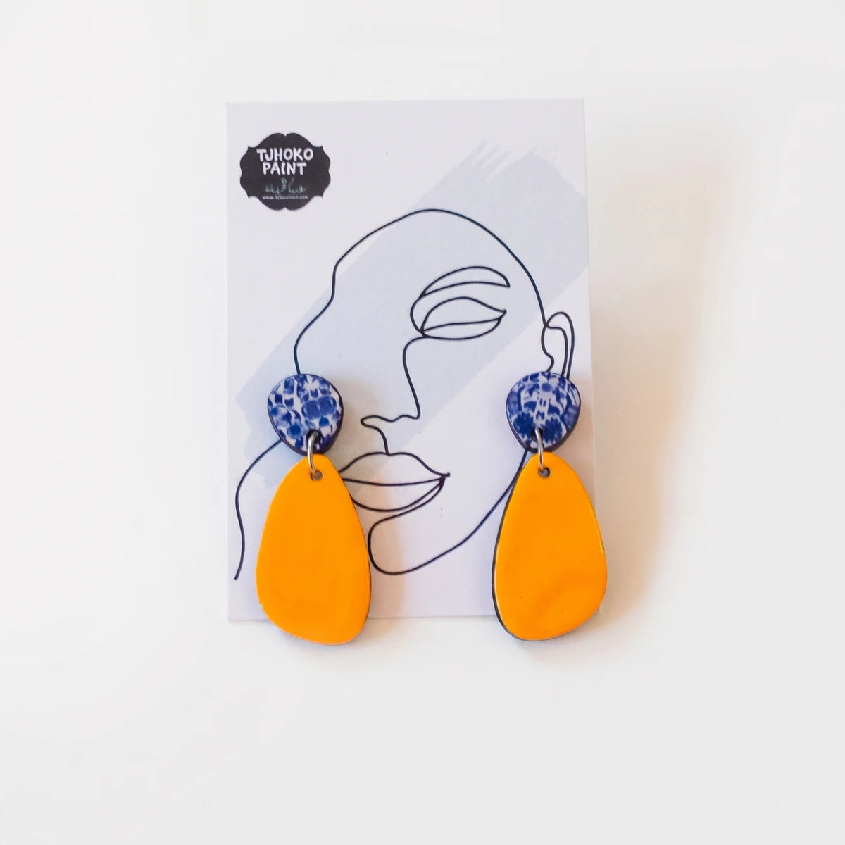 A pair of Tjhoko John's Delft earrings featuring orange teardrop dangles and blue circular studs with white patterns,