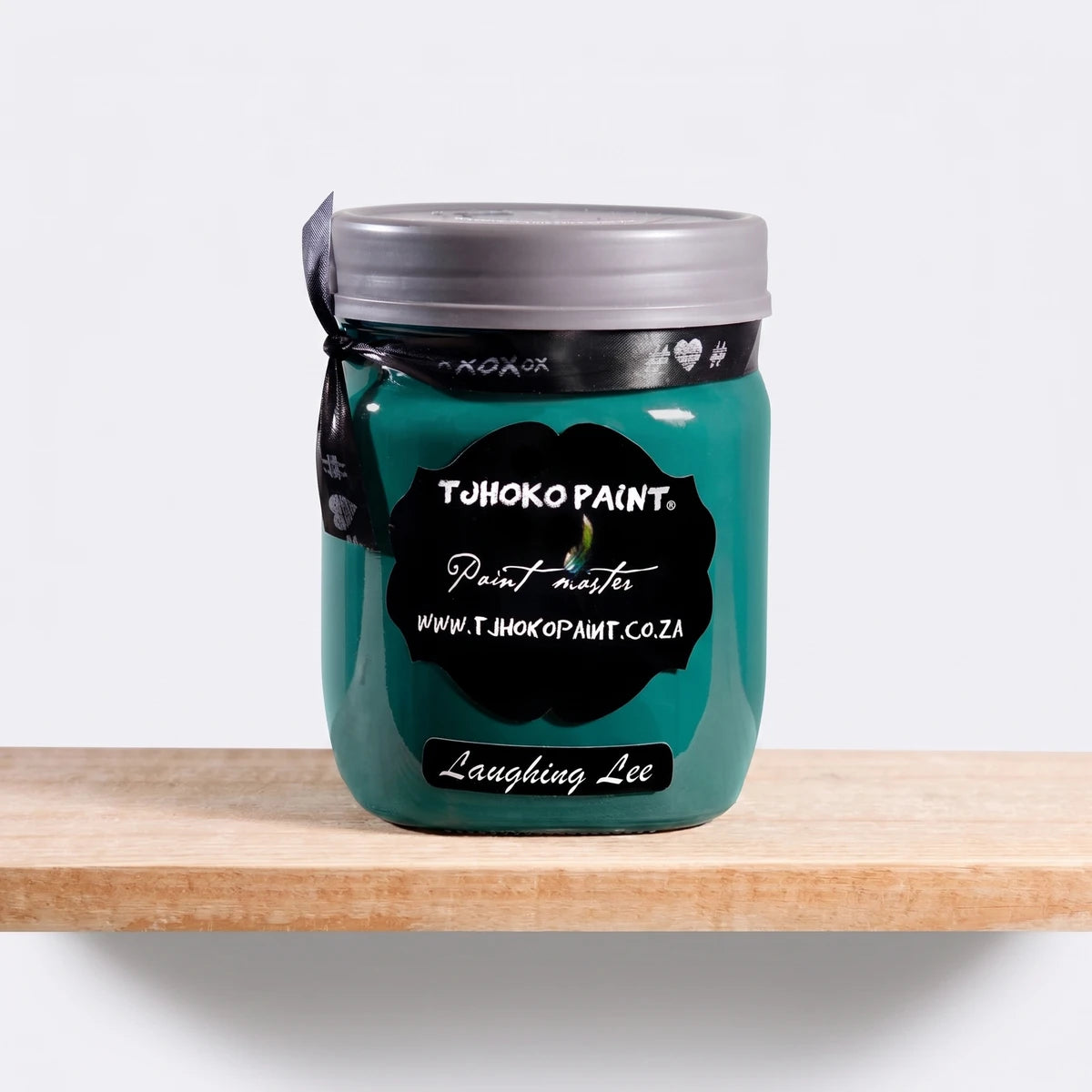 Tjhoko Paint Laughing Lee 500ml chalk paint in a teal glass jar with a grey lid and black ribbon, on a light wooden shelf.