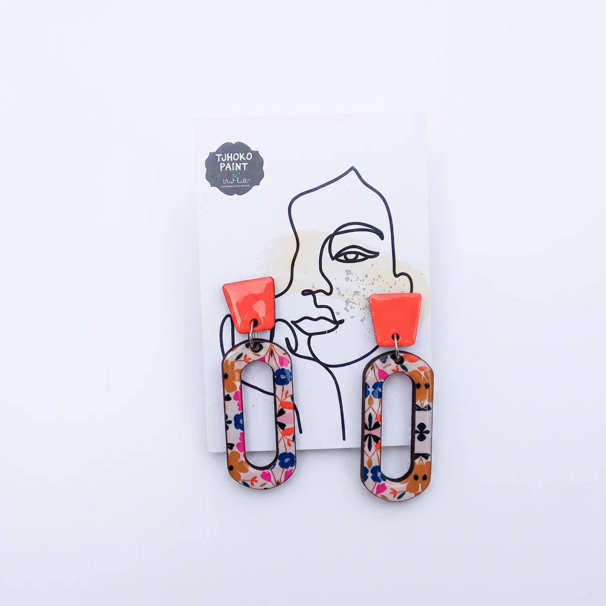 Tjhoko Paint P's Flower Power Earrings feature a vibrant floral pattern on an oval dangle with a solid coral top, displayed
