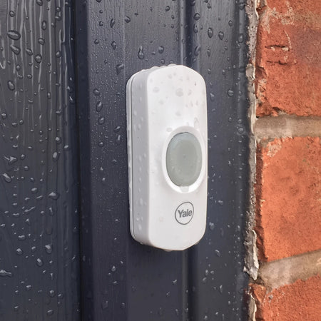 White Yale wirefree doorbell chime, wet with rain droplets, mounted on a dark gray door.